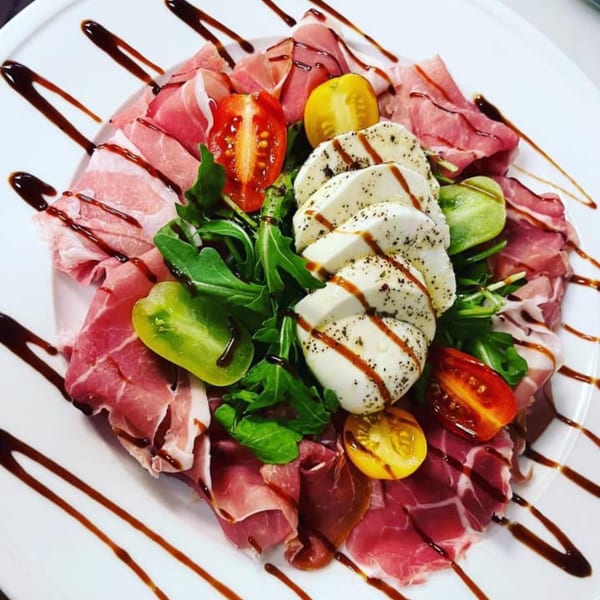 A white plate with prosciutto, fresh mozzarella, cherry tomatoes, arugula, and balsamic glaze.