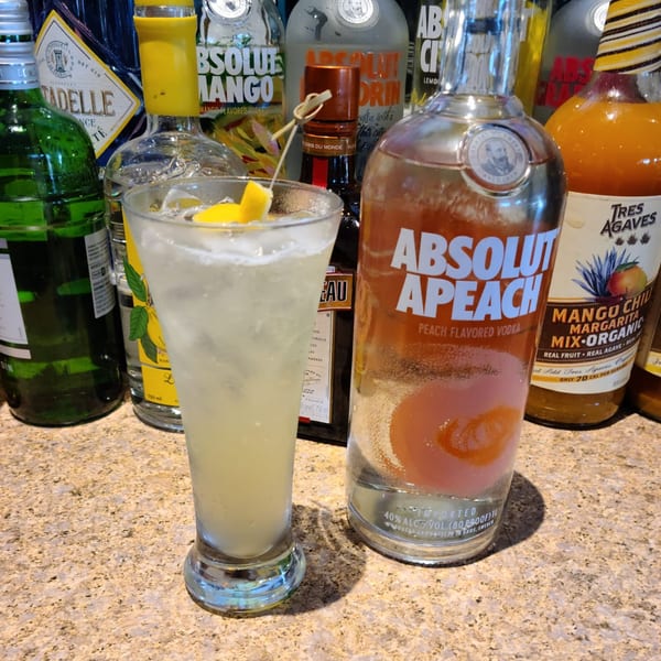 A tall glass of light-colored cocktail with lemon wedges and ice, surrounded by liquor bottles.