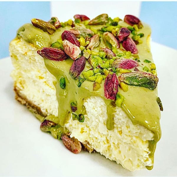 Slice of cheesecake topped with green pistachio sauce and whole pistachios on a white plate.