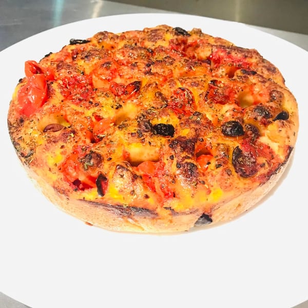 Round focaccia bread topped with cherry tomatoes, black olives, and herbs on a white plate.
